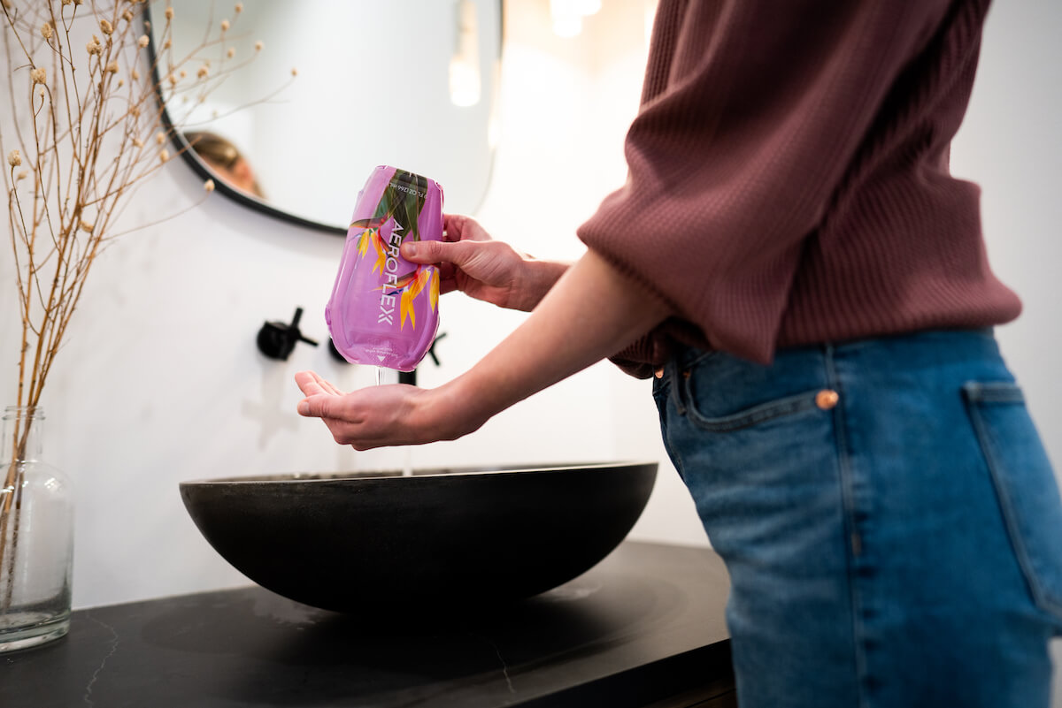 Woman pouring liquid from a purple pouch AeroFlexx pouch into their hand over a black sink.