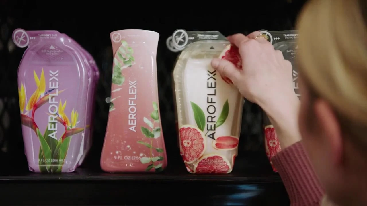 A person picking up a cream-colored AeroFlexx pouch with a pomegranate design from a shelf with three drink pouches.